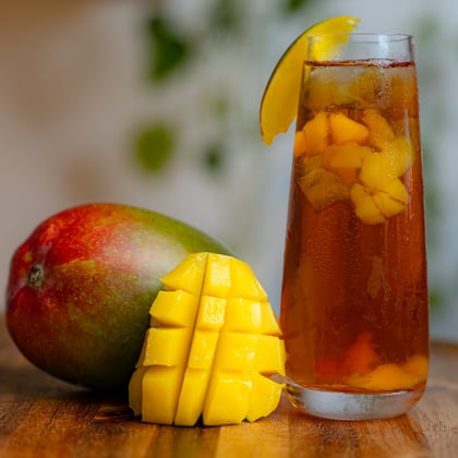 A bright cut mango sitting on a wooden table next to an ice cold glass of Firepot mango bergamot iced tea