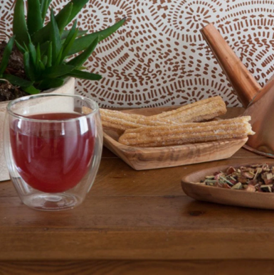 red hibiscus elixir in a clear cup on wooden table next to loose leaf tea and a green plant