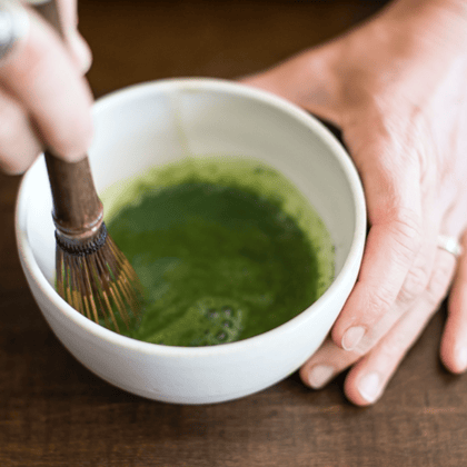liquid matcha in a small white cup being stirred by a wooden matcha whisk